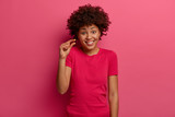 Positive African American woman shows something very small or tiny, discusses prices in store, smiles happily, dressed casually, says little bit, isolated over pink background. Size concept.