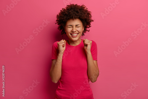 Ecstatic joyful woman makes fist bump, feels very happy, rejoices success and triumph, celebrates much money in lotery, closes eyes, dressed in pink clothes, poses indoor. Victory accomplished