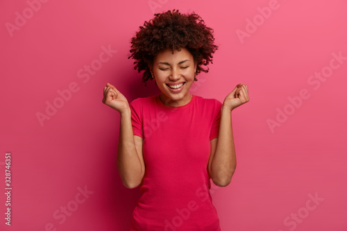 Image of happy woman makes fist pump gesture, celebrates great news, giggles positively, wears casual clothes, laughs positively, poses against bright pink background. Emotions and success concept