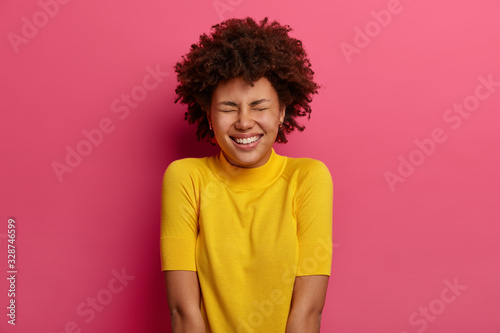 Positive African American woman smiles happily, laughs at funny joke, wears yellow clothes, closes eyes with pleasure, isolated over pink background. People, emotions and face expressions concept