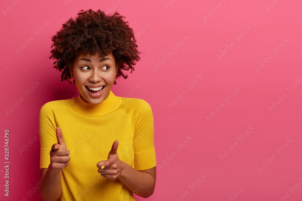 Positive dark skinned young female points at camera, chooses you, feels happy and relaxed, giggles gladfully, looks aside, wears yellow t shirt, poses against pink studio wall, copy space aside
