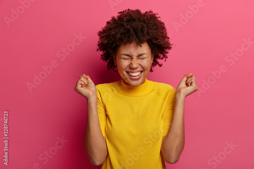 Cheerful curly haired woman clenches fists with positive expression, rejoices wonderful news, smiles happily, squints face with pleasure, wears yellow clothes, stands against pink vibrant background