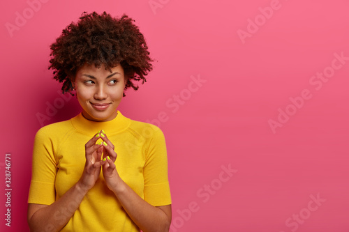 My plan is perfect. Pretty African American woman schemes something, steepls fingers and looks with cunning expression aside, smiles sly, poses over pink wall, copy space aside for your promo