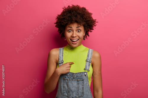 Joyful surprised African American woman indicates at herself, asks really me, has positive expression, unexpected to hear something, wears denim overalls stands indoor shocked being picked or selected