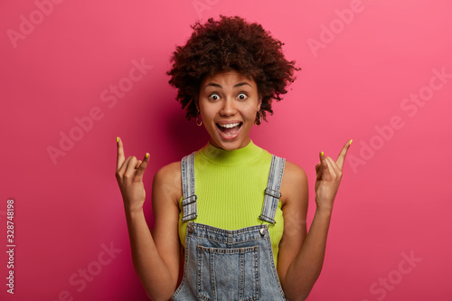 Excited cheerful woman born to be rock star, shows horn hand gesture, enjoys punk rock music, stays wild and free, being heavy metal fan, dressed in fashionable clothes, poses indoor over rosy wall