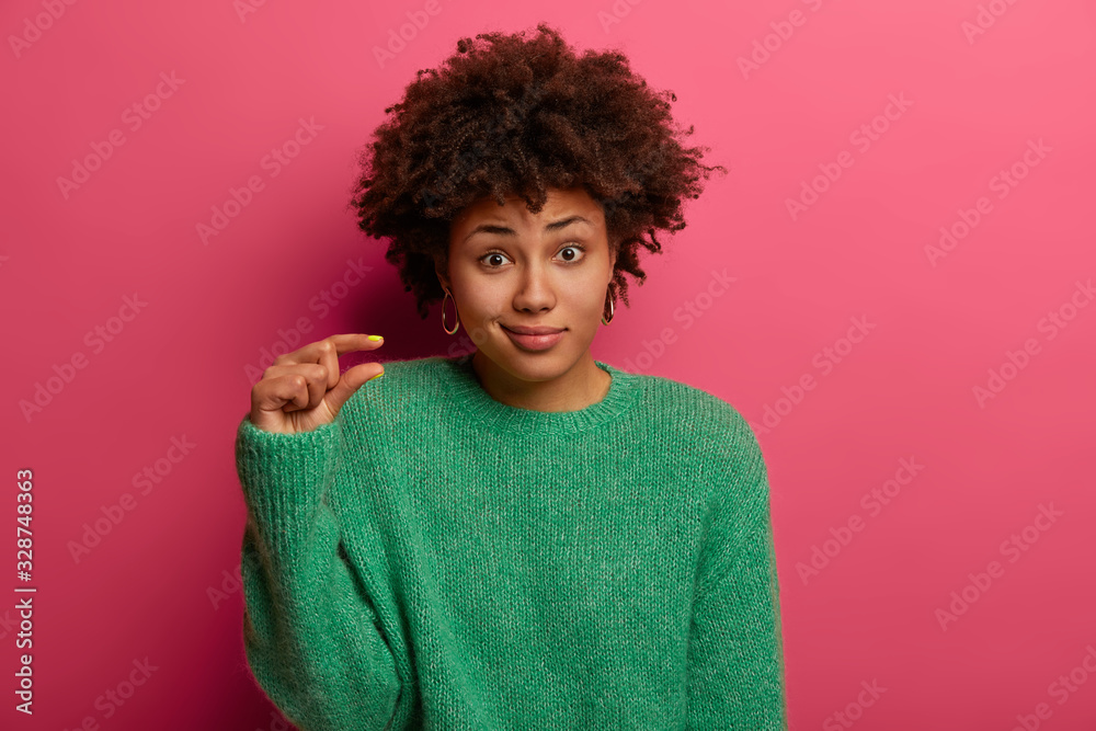 Curly woman measures something tiny, says she has little problems, shapes small obejct, wears green sweater, looks unimpressed at camera, poses over rosy background. Body language and size concept Curly woman measures something tiny, says she has little problems, shapes small obejct, wears green sweater, looks unimpressed at camera, poses over rosy background. Body language and size concept