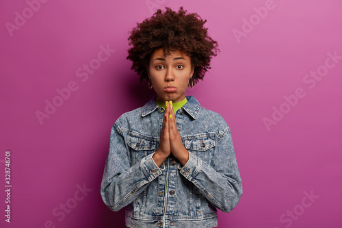 Innocent dark skinned woman keeps palms pressed together, looks with imploring sad expression, pleads for something, wears denim jacket, poses against purple background. Please, help me once again © Wayhome Studio