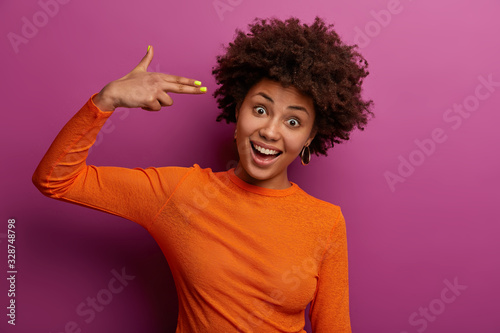Horizontal shot of positive curly woman shoots in temples, makes suicide gesture, foolishes around, makes finger gun pistol, laughs happily, wears orange jumper, isolated on purple background