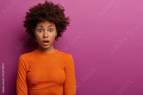 Canvastavla Indoor shot of shocked curly haired woman horrified to find out about awful accident, dressed in orange jumper, stands against purple background, surprised by sudden news