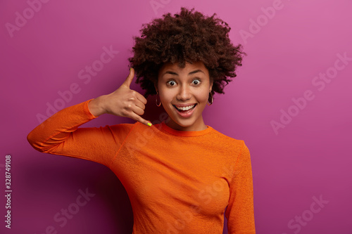 Call me later. Pleased curly young woman makes phone gesture, pretends mobile communication, smiles happily, wears casual orange jumper, isolated over purple studio wall. Body language concept © wayhome.studio 