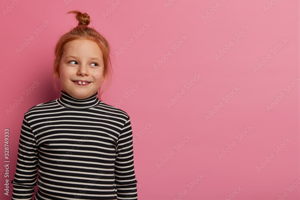 Studio shot of ginger girl looks gladfully aside, has two teeth stick out, wears striped jumper, being in good mood after coming home from kindergarten, poses indoor over rosy wall. Children concept Studio shot of ginger girl looks gladfully aside, has two teeth stick out, wears striped jumper, being in good mood after coming home from kindergarten, poses indoor over rosy wall. Children concept