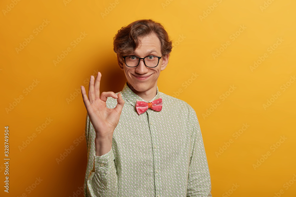 Funny smiling male geek shows ring gesture with fingers, says good job, satisfied with suggestion, wears spectacles and formal shirt, looks gladfully at camera, isolated on yellow background - obrazy, fototapety, plakaty 
