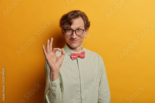 Funny smiling male geek shows ring gesture with fingers, says good job, satisfied with suggestion, wears spectacles and formal shirt, looks gladfully at camera, isolated on yellow background photo