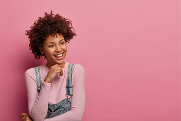 Portrait of cheerful tender young woman with curly Afro hair smiles gently and looks pensively aside, has charming expression, thinks about pleasant moment in life, isolated on pink background