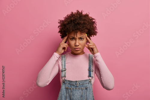 Young beautiful tired woman suffers migraine or headache  touches temples and looks intense  asks for painkillers  wears denim sarafan  stands against pink pastel background. Negative feelings