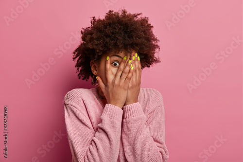 Frightened woman covers face with palms, peeks through fingers, sees something horrible and unexpected, hides from all difficulties, intrigued what happening, wears jumper in one tone with background photo
