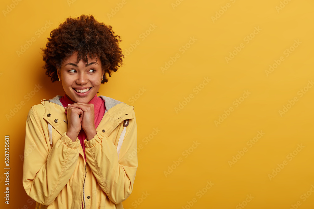 Smiling tender young woman keeps hands under chin, gazes aside with broad toothy smile, wears casual anorak, dreams about something pleasant, poses against yellow studio wall, empty space area Smiling tender young woman keeps hands under chin, gazes aside with broad toothy smile, wears casual anorak, dreams about something pleasant, poses against yellow studio wall, empty space area