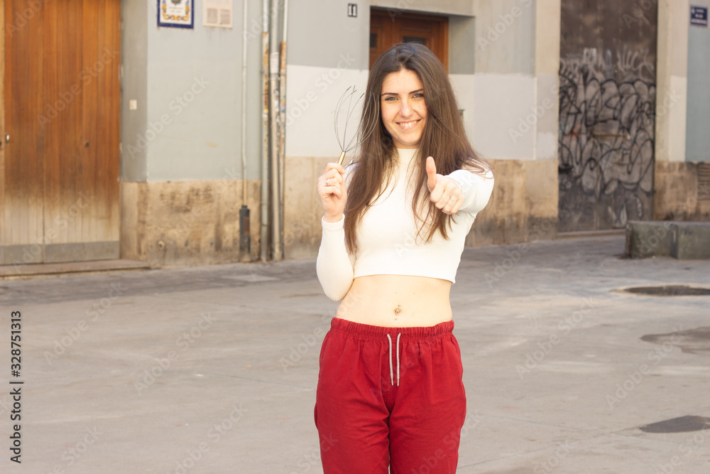 Portrait of pretty young woman with neural massager  showing thumb up gesture, in the city, white top and red pants, dark air. Place for your text in copy space.