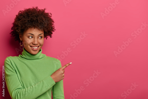 Horizontal shot of smiling curly haired woman indicates at free space, demonstrates place for your advertisement, attracts attention to sale, wears green turtleneck, isolated on vibrant pink wall