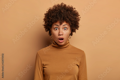 Headshot of stunned woman with Afro hair, wonders something, gasps from excitement, keeps mouth opened, dressed in casual poloneck, isolated over beige background. No way. Unlikely to be true