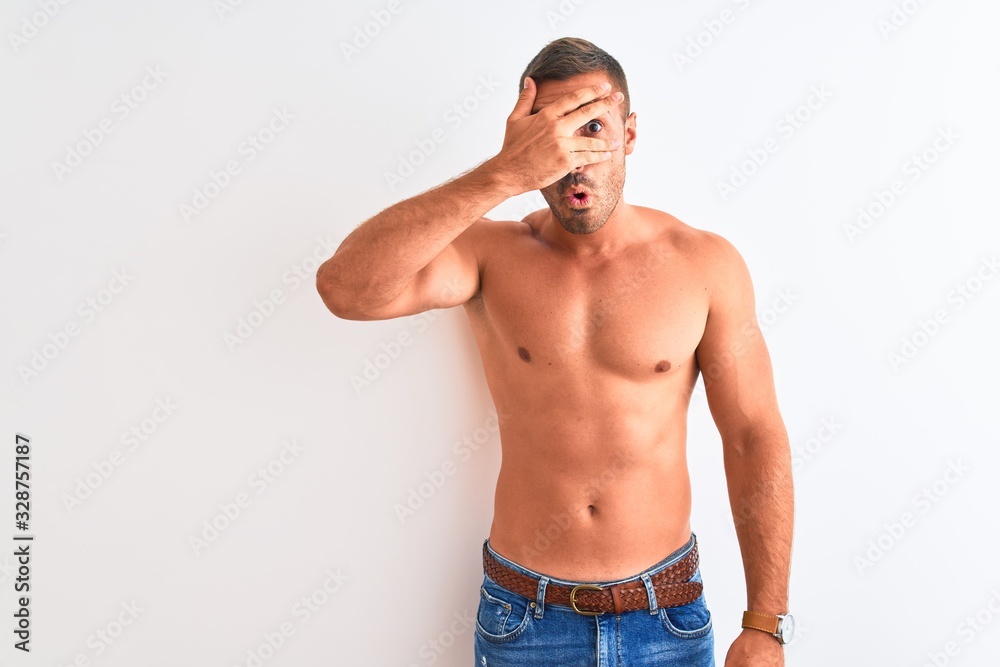 Young handsome shirtless man showing muscular body over isolated background peeking in shock covering face and eyes with hand, looking through fingers with embarrassed expression.