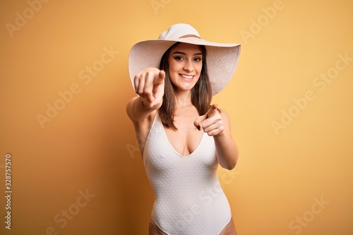 Young beautiful brunette woman on vacation wearing swimsuit and summer hat pointing to you and the camera with fingers, smiling positive and cheerful