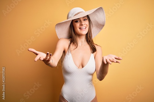 Young beautiful brunette woman on vacation wearing swimsuit and summer hat smiling cheerful offering hands giving assistance and acceptance.