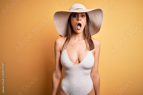 Young beautiful brunette woman on vacation wearing swimsuit and summer hat afraid and shocked with surprise and amazed expression, fear and excited face.