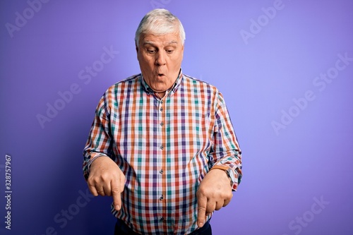 Senior handsome hoary man wearing casual colorful shirt over isolated purple background Pointing down with fingers showing advertisement, surprised face and open mouth