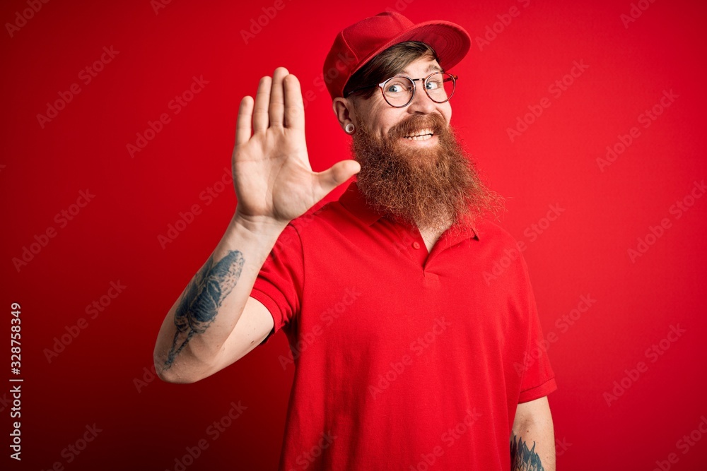 Naklejka premium Young handsome delivery man wearing glasses and red cap over isolated background Waiving saying hello happy and smiling, friendly welcome gesture
