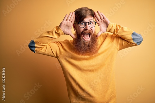 Handsome Irish redhead man with beard wearing glasses over yellow isolated background Smiling cheerful playing peek a boo with hands showing face. Surprised and exited photo