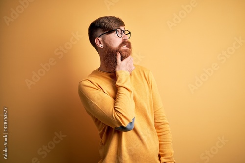 Handsome Irish redhead man with beard wearing glasses over yellow isolated background Thinking worried about a question  concerned and nervous with hand on chin