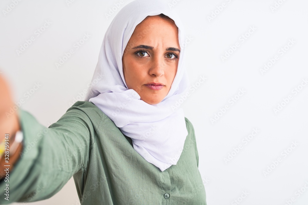 Young Arab woman wearing hijab taking selfie photo over isolated background with a confident expression on smart face thinking serious Young Arab woman wearing hijab taking selfie photo over isolated background with a confident expression on smart face thinking serious