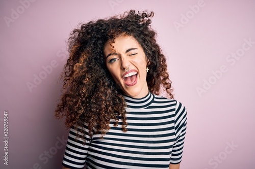 Young beautiful woman with curly hair and piercing wearing casual striped t-shirt winking looking at the camera with sexy expression, cheerful and happy face.