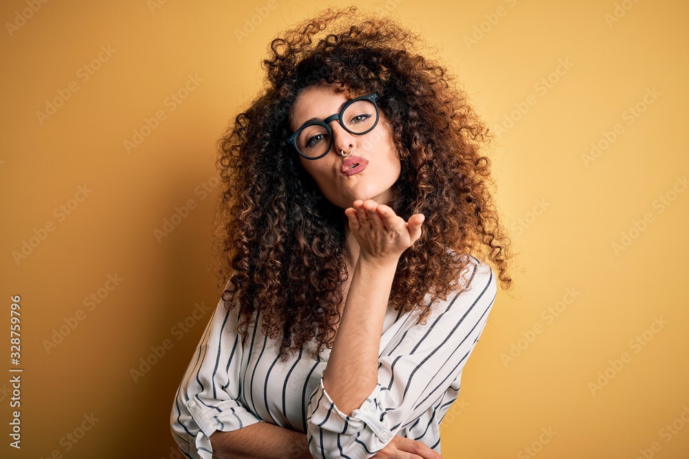 Young beautiful woman with curly hair and piercing wearing striped shirt and glasses looking at the camera blowing a kiss with hand on air being lovely and sexy. Love expression. Young beautiful woman with curly hair and piercing wearing striped shirt and glasses looking at the camera blowing a kiss with hand on air being lovely and sexy. Love expression.