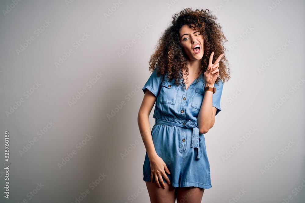 Young beautiful woman with curly hair and piercing wearing casual denim dress smiling with happy face winking at the camera doing victory sign. Number two. Young beautiful woman with curly hair and piercing wearing casual denim dress smiling with happy face winking at the camera doing victory sign. Number two.