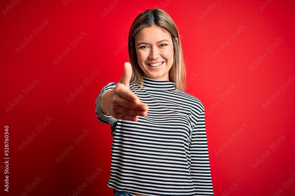 Young beautiful blonde woman wearing casual striped sweater over red isolated background smiling friendly offering handshake as greeting and welcoming. Successful business.