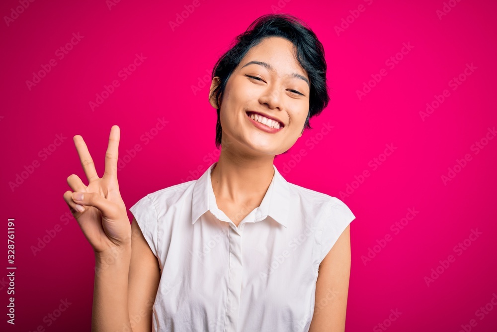 Young beautiful asian girl wearing casual summer shirt standing over isolated pink background smiling with happy face winking at the camera doing victory sign. Number two. Young beautiful asian girl wearing casual summer shirt standing over isolated pink background smiling with happy face winking at the camera doing victory sign. Number two.
