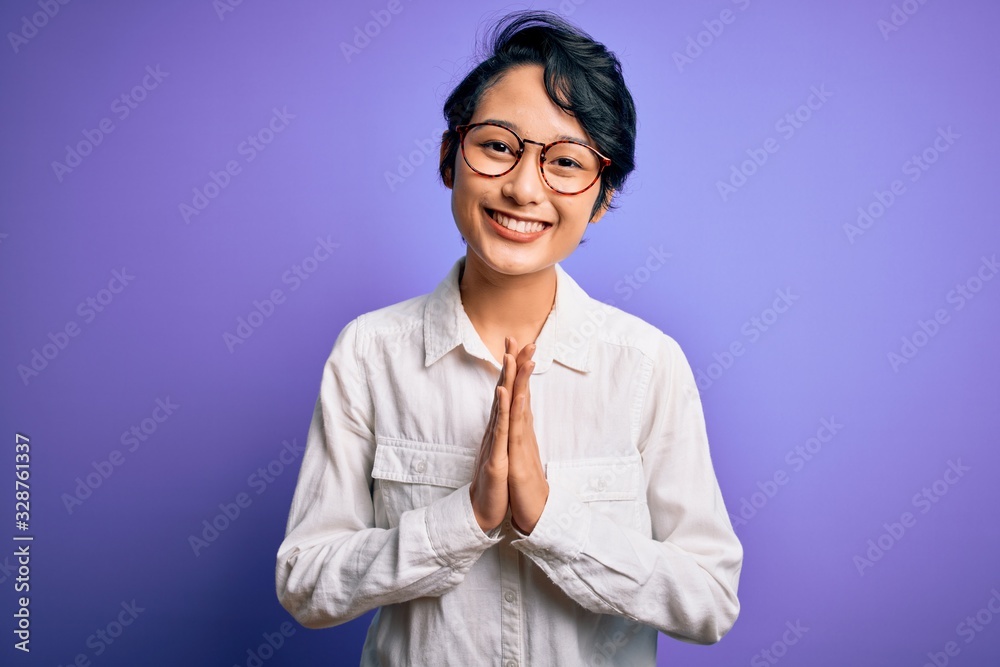 Young beautiful asian girl wearing casual shirt and glasses standing over purple background praying with hands together asking for forgiveness smiling confident. Young beautiful asian girl wearing casual shirt and glasses standing over purple background praying with hands together asking for forgiveness smiling confident.