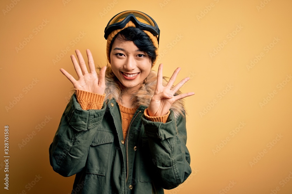 Beautiful asian skier girl wearing snow sportswear using ski goggles over yellow background showing and pointing up with fingers number nine while smiling confident and happy. Beautiful asian skier girl wearing snow sportswear using ski goggles over yellow background showing and pointing up with fingers number nine while smiling confident and happy.