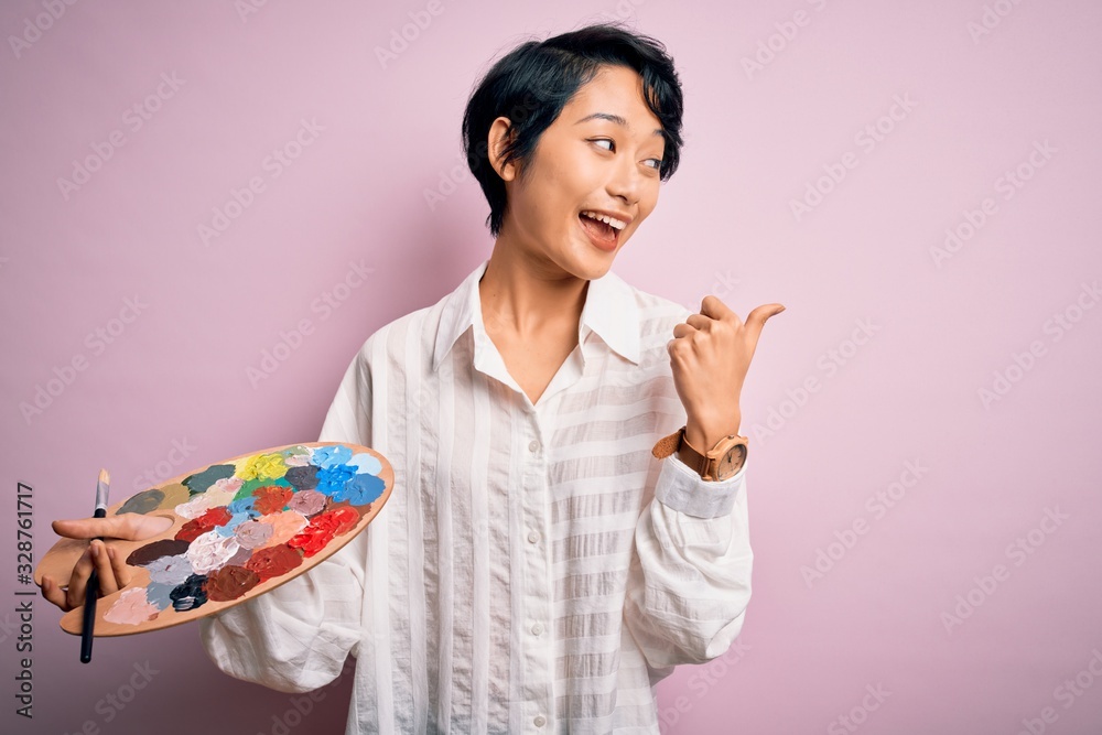 Young beautiful chinese artist woman painting using paintbrush and palette with colors pointing and showing with thumb up to the side with happy face smiling