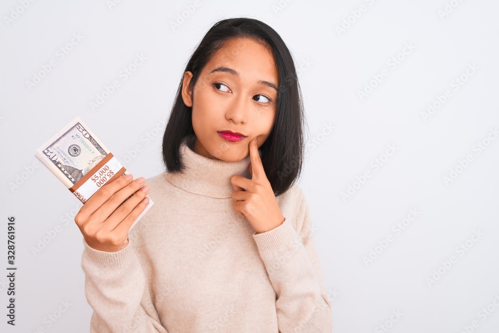 Young beautiful chinese woman holding dollars standing over isolated white background serious face thinking about question, very confused idea Young beautiful chinese woman holding dollars standing over isolated white background serious face thinking about question, very confused idea
