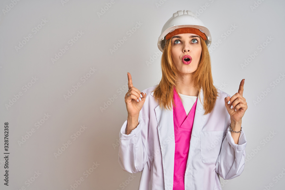 Redhead caucasian woman engineer wearing safety helmet over isolated background amazed and surprised looking up and pointing with fingers and raised arms. Redhead caucasian woman engineer wearing safety helmet over isolated background amazed and surprised looking up and pointing with fingers and raised arms.