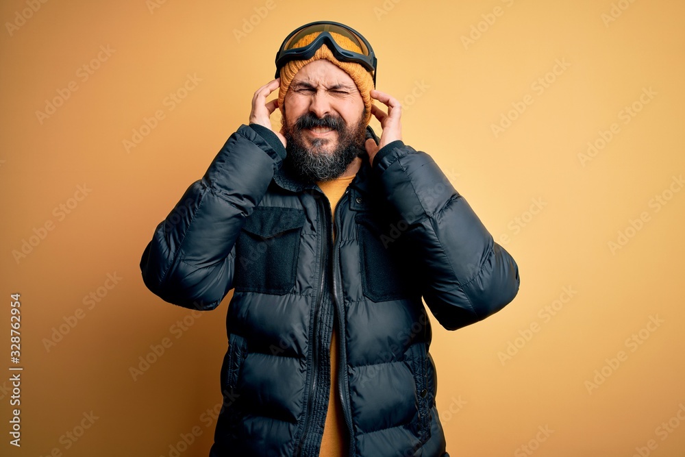 Handsome skier bald man with beard skiing wearing snow sportswear and ski goggles covering ears with fingers with annoyed expression for the noise of loud music. Deaf concept.