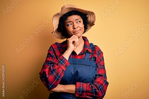 Young African American afro farmer woman with curly hair wearing apron and hat with hand on chin thinking about question, pensive expression. Smiling with thoughtful face. Doubt concept.