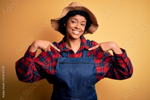 Young African American afro farmer woman with curly hair wearing apron and hat looking confident with smile on face, pointing oneself with fingers proud and happy.