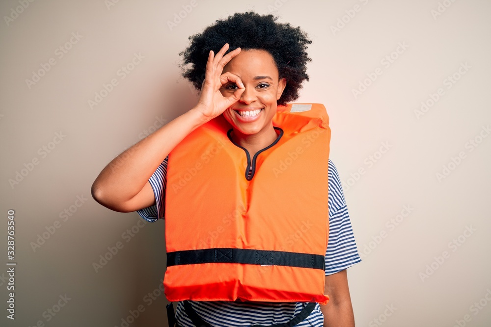 Young African American afro woman with curly hair wearing orange protection lifejacket doing ok gesture with hand smiling, eye looking through fingers with happy face. Young African American afro woman with curly hair wearing orange protection lifejacket doing ok gesture with hand smiling, eye looking through fingers with happy face.