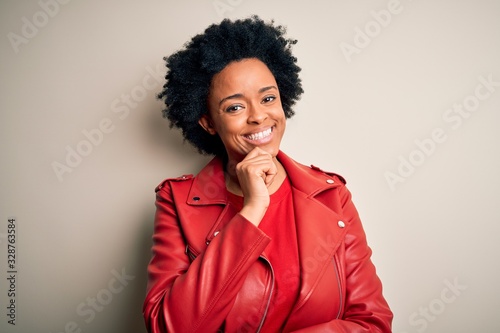 Young beautiful African American afro woman with curly hair wearing casual red jacket looking confident at the camera with smile with crossed arms and hand raised on chin. Thinking positive.