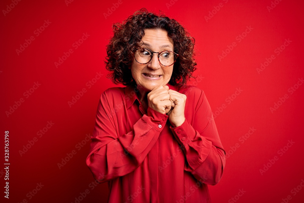 Middle age beautiful curly hair woman wearing casual shirt and glasses over red background laughing nervous and excited with hands on chin looking to the side Middle age beautiful curly hair woman wearing casual shirt and glasses over red background laughing nervous and excited with hands on chin looking to the side