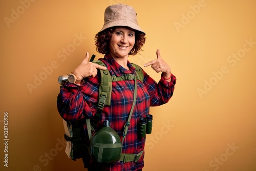 Middle age curly hair hiker woman hiking wearing backpack and water canteen using binoculars looking confident with smile on face, pointing oneself with fingers proud and happy.